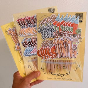 May include: Multiple sets of decorative paper cupcake toppers attached to wooden picks, packaged on yellow cards. The designs include zebras, giraffes, lions, the number one, and a child's face, with the text Cuspper - Tiny Toppers, Big Celebrations.