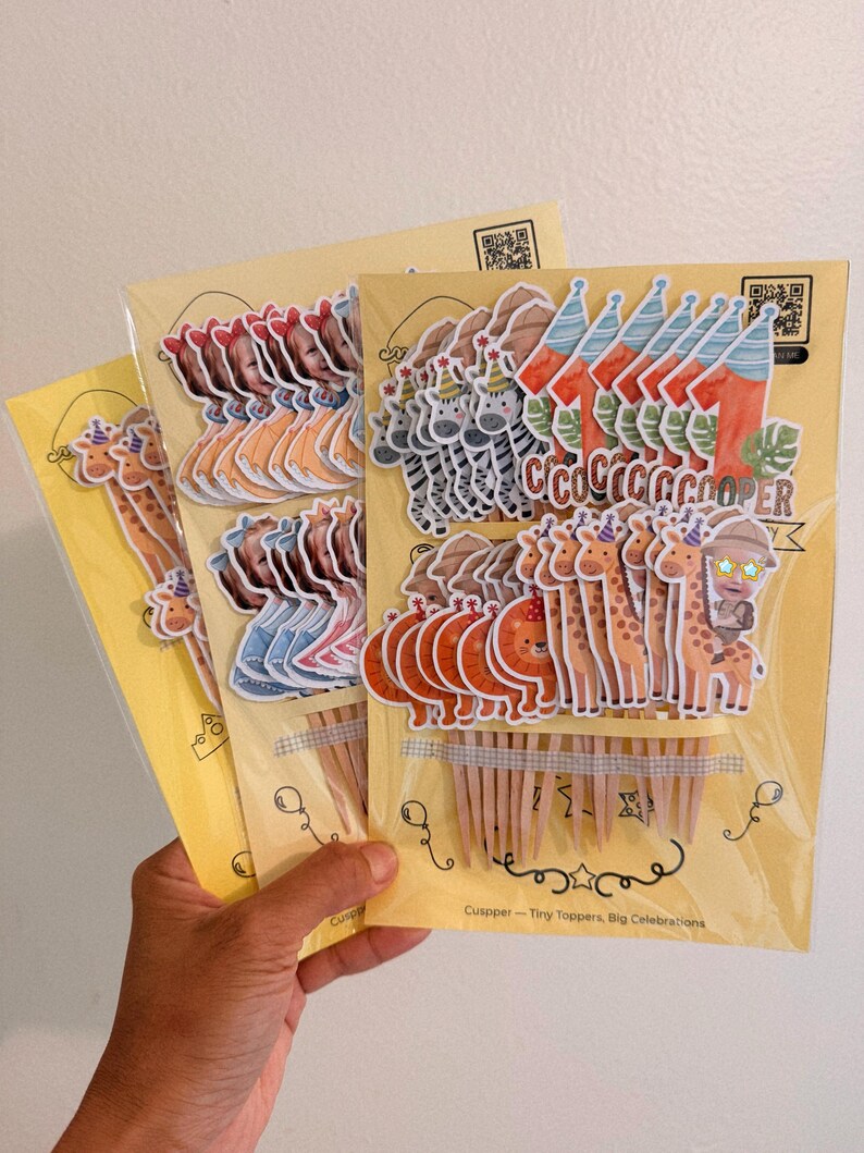 May include: Multiple sets of decorative paper cupcake toppers attached to wooden picks, packaged on yellow cards. The designs include zebras, giraffes, lions, the number one, and a child's face, with the text Cuspper - Tiny Toppers, Big Celebrations.