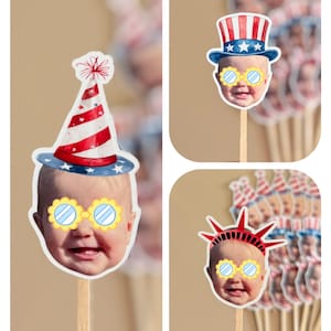 May include: A collection of custom cupcake toppers featuring a baby's face wearing yellow-rimmed glasses. The toppers include illustrations of a red and white striped party hat, a patriotic top hat, and a red Statue of Liberty crown on wooden sticks.