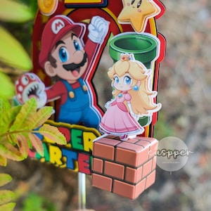 Peut inclure: D&eacute;coration de g&acirc;teau color&eacute;e avec Mario, la Princesse Peach et d'autres personnages de Super Mario Bros. La d&eacute;coration comprend un grand chiffre 1, une &eacute;toile, une pi&egrave;ce et les mots "Super Mario" dans un design vibrant. Les personnages sont sur fond rouge et jaune.