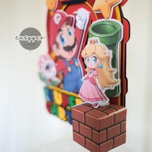 Peut inclure: D&eacute;coration de g&acirc;teau sur le th&egrave;me de Super Mario, avec Mario, la Princesse Peach et un tuyau vert. La d&eacute;coration comprend un bloc de briques et une &eacute;toile, avec un code couleur rouge, jaune et bleu. Le texte "cuspper" est visible.