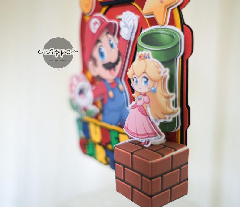 Peut inclure: D&eacute;coration de g&acirc;teau sur le th&egrave;me de Super Mario, avec Mario, la Princesse Peach et un tuyau vert. La d&eacute;coration comprend un bloc de briques et une &eacute;toile, avec un code couleur rouge, jaune et bleu. Le texte "cuspper" est visible.