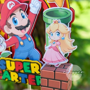 Peut inclure: D&eacute;coration de g&acirc;teau sur le th&egrave;me de Super Mario avec Mario, la princesse Peach, un tuyau vert et une &eacute;toile. Le d&eacute;cor comprend les mots "SUPER ARTER" en jaune et vert.