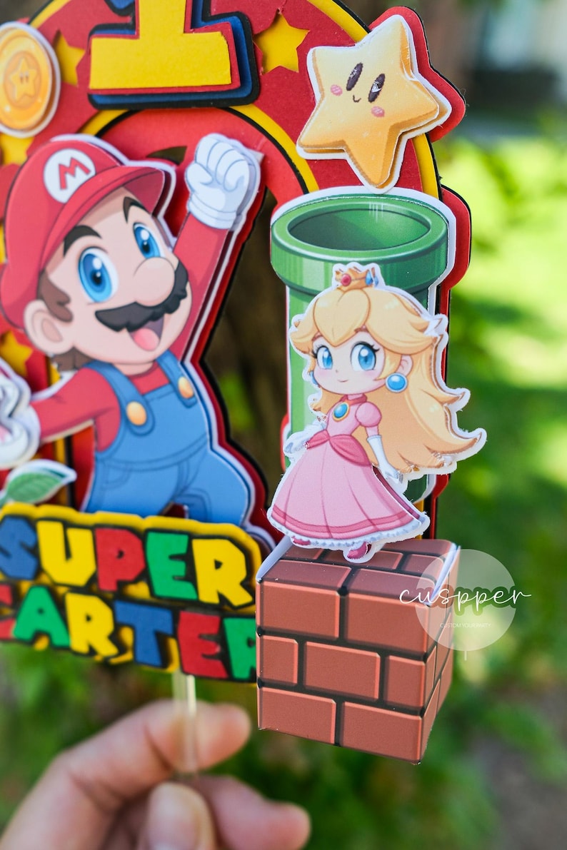 Peut inclure: D&eacute;coration de g&acirc;teau sur le th&egrave;me de Super Mario avec Mario, la princesse Peach, un tuyau vert et une &eacute;toile. Le d&eacute;cor comprend les mots "SUPER ARTER" en jaune et vert.