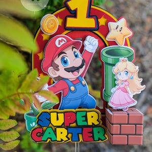Peut inclure: D&eacute;coration de g&acirc;teau sur le th&egrave;me de Super Mario, avec Mario, la Princesse Peach, un tuyau vert et une &eacute;toile. Le chiffre 1 est affich&eacute; en jaune, et le nom "SUPER CARTER" est &eacute;crit en lettres color&eacute;es. Une pi&egrave;ce est &eacute;galement incluse.
