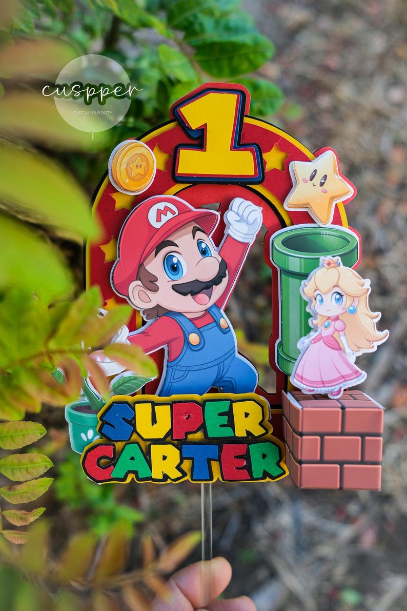 Peut inclure: D&eacute;coration de g&acirc;teau sur le th&egrave;me de Super Mario, avec Mario, la Princesse Peach, un tuyau vert et une &eacute;toile. Le chiffre 1 est affich&eacute; en jaune, et le nom "SUPER CARTER" est &eacute;crit en lettres color&eacute;es. Une pi&egrave;ce est &eacute;galement incluse.
