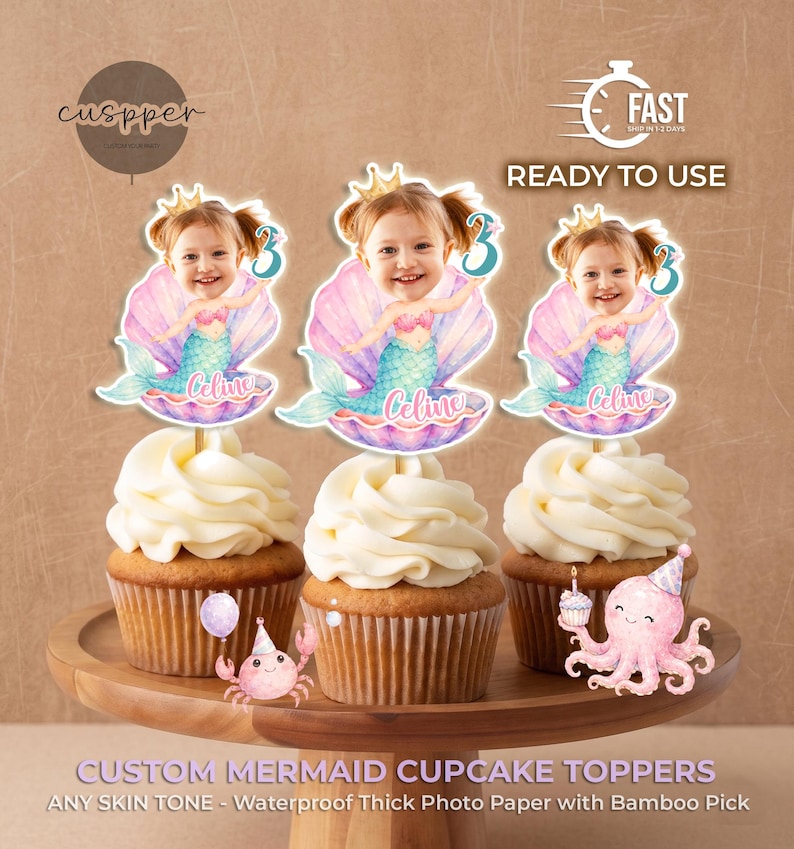 May include: Three cupcakes topped with custom mermaid decorations featuring a child's face, a gold crown, a teal tail, and a pink seashell. The name Celine is printed in script, along with the number 3. Includes a small pink crab and octopus.