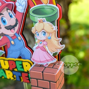 Peut inclure: D&eacute;coration de g&acirc;teau sur le th&egrave;me de Super Mario, avec la princesse Peach en robe rose sur un bloc de briques. Mario, un tuyau vert et une &eacute;toile sont &eacute;galement pr&eacute;sents. Le texte "SUPER ARTE" est visible.