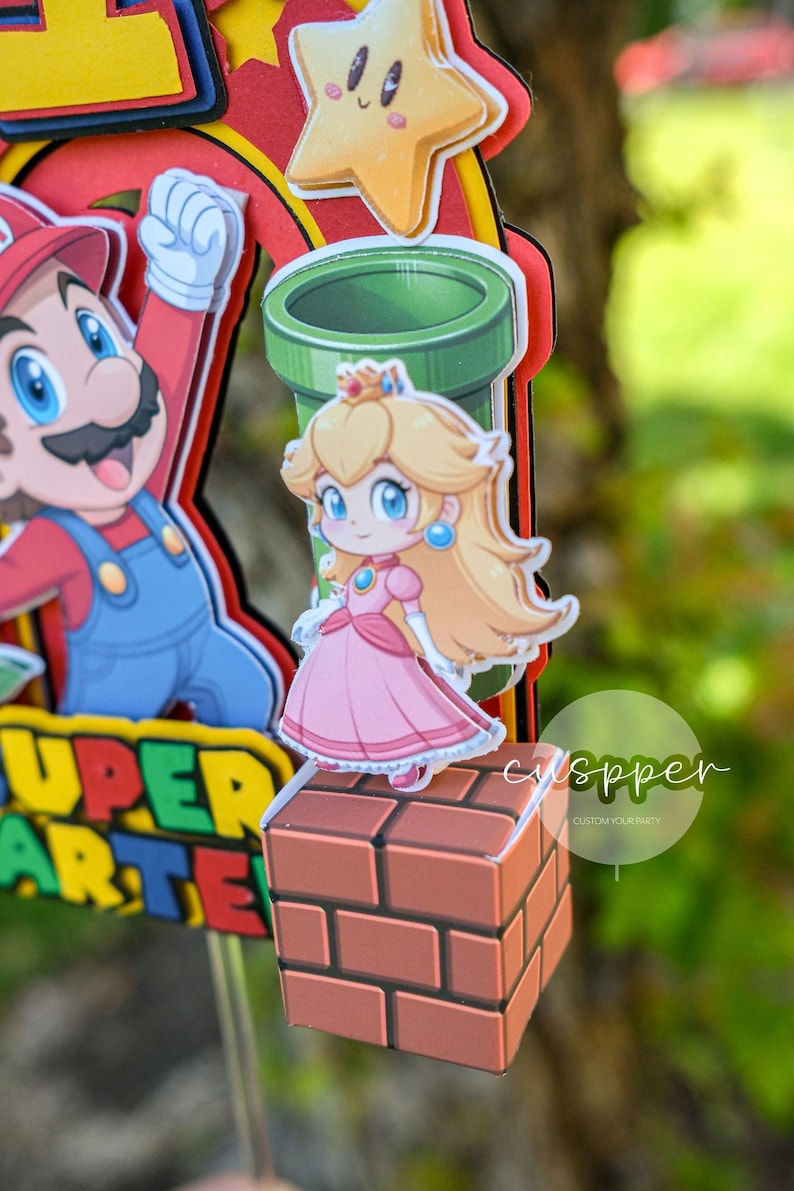 Peut inclure: D&eacute;coration de g&acirc;teau sur le th&egrave;me de Super Mario, avec la princesse Peach en robe rose sur un bloc de briques. Mario, un tuyau vert et une &eacute;toile sont &eacute;galement pr&eacute;sents. Le texte "SUPER ARTE" est visible.