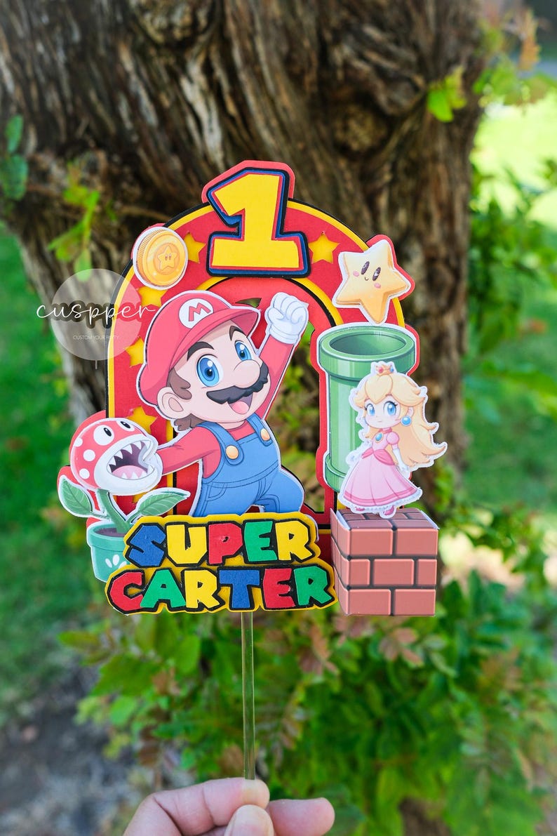 Peut inclure: D&eacute;coration de g&acirc;teau sur le th&egrave;me de Super Mario avec Mario, la Princesse Peach, une plante Piranha et un tuyau vert. Le chiffre 1 est mis en &eacute;vidence, avec le nom "SUPER CARTER". Parfait pour une f&ecirc;te d'anniversaire.