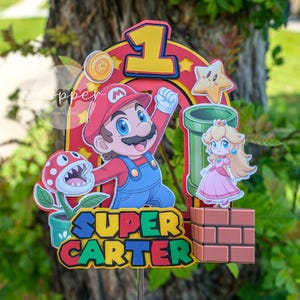 Peut inclure: D&eacute;coration de g&acirc;teau sur le th&egrave;me de Super Mario avec Mario, la Princesse Peach, un tuyau vert et une plante Piranha. Le topper comprend le chiffre 1 et le nom "Carter" dans une police ludique, parfait pour un anniversaire.