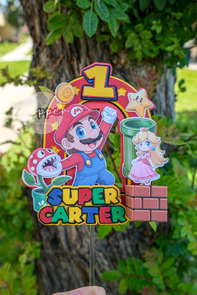 Peut inclure: D&eacute;coration de g&acirc;teau sur le th&egrave;me de Super Mario avec Mario, la Princesse Peach, un tuyau vert et une plante Piranha. Le topper comprend le chiffre 1 et le nom "Carter" dans une police ludique, parfait pour un anniversaire.