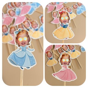 May include: A collection of custom cupcake toppers featuring a child's face cutout attached to a wooden stick. Each topper depicts a cartoon princess body in various colors including light blue, yellow, and pink, with yellow flower-shaped glasses.