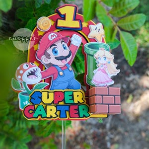 Peut inclure: D&eacute;coration de g&acirc;teau sur le th&egrave;me de Super Mario, avec Mario, la Princesse Peach, une plante Piranha et un tuyau vert. Le dessus de g&acirc;teau comprend le chiffre 1 et le nom "SUPER CARTER". Le design est plac&eacute; sur un fond de feuillage vert.