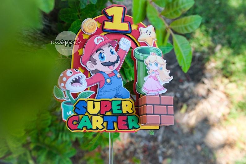 Peut inclure: D&eacute;coration de g&acirc;teau sur le th&egrave;me de Super Mario, avec Mario, la Princesse Peach, une plante Piranha et un tuyau vert. Le dessus de g&acirc;teau comprend le chiffre 1 et le nom "SUPER CARTER". Le design est plac&eacute; sur un fond de feuillage vert.