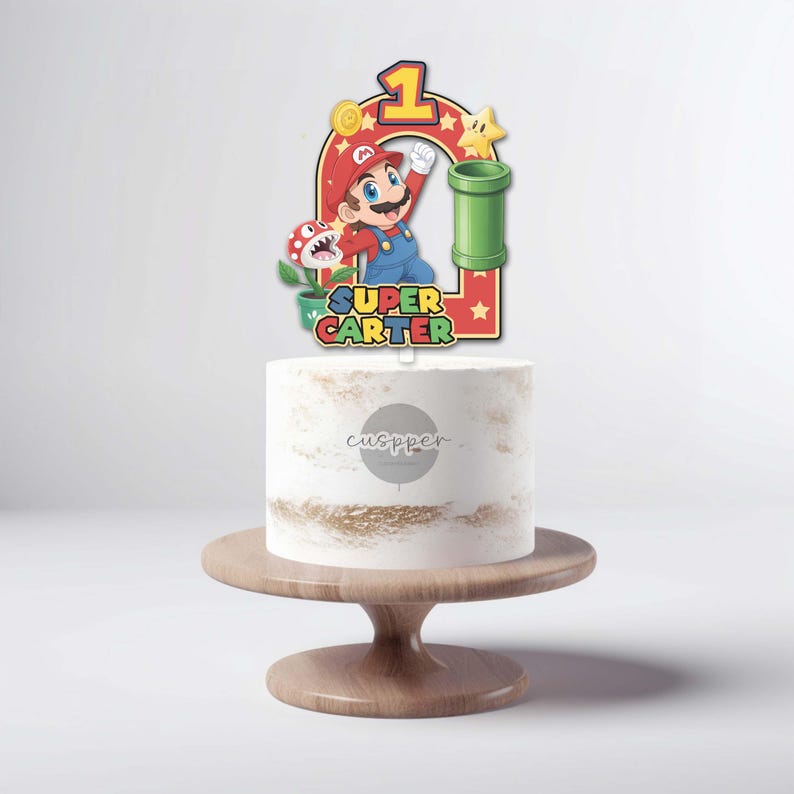 Peut inclure: Un cake topper color&eacute; sur le th&egrave;me de Super Mario Bros. avec Mario, un tuyau vert et le chiffre 1. Le texte indique "Super Carter". Le g&acirc;teau est blanc avec des d&eacute;tails dor&eacute;s, pr&eacute;sent&eacute; sur un pr&eacute;sentoir &agrave; g&acirc;teau en bois.