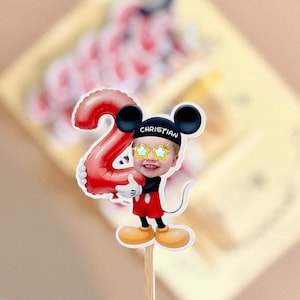 May include: A cake topper featuring a cartoon mouse character wearing a black hat with the name CHRISTIAN printed in white, yellow star-shaped glasses, and red shorts, holding a large red balloon shaped like the number two on a wooden stick.
