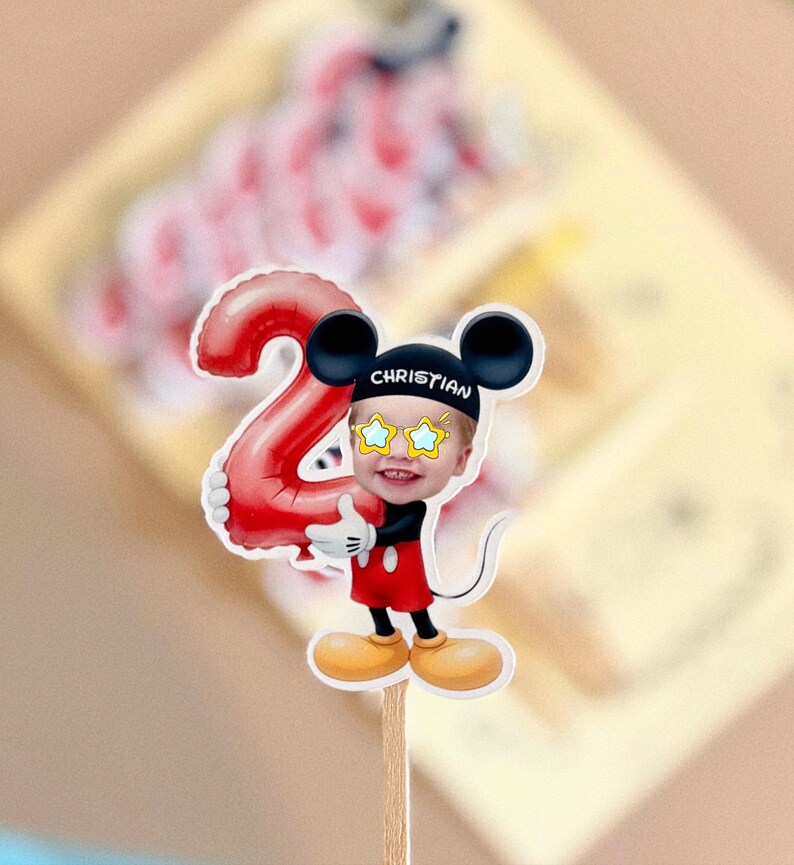 May include: A cake topper featuring a cartoon mouse character wearing a black hat with the name CHRISTIAN printed in white, yellow star-shaped glasses, and red shorts, holding a large red balloon shaped like the number two on a wooden stick.