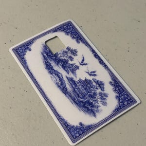 May include: A rectangular, white and blue decorative card with a landscape design. The card features a blue and white illustration of a scenic landscape with birds and trees. The edges are trimmed with a blue floral pattern and a square cut-out.