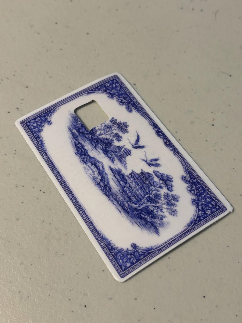 May include: A rectangular, white and blue decorative card with a landscape design. The card features a blue and white illustration of a scenic landscape with birds and trees. The edges are trimmed with a blue floral pattern and a square cut-out.