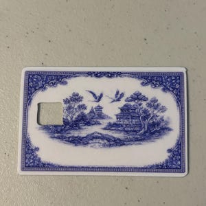 May include: A rectangular, white and blue card with a decorative design. The blue design features a landscape with buildings, trees, and flying birds. The card has a rectangular cutout on the left side.