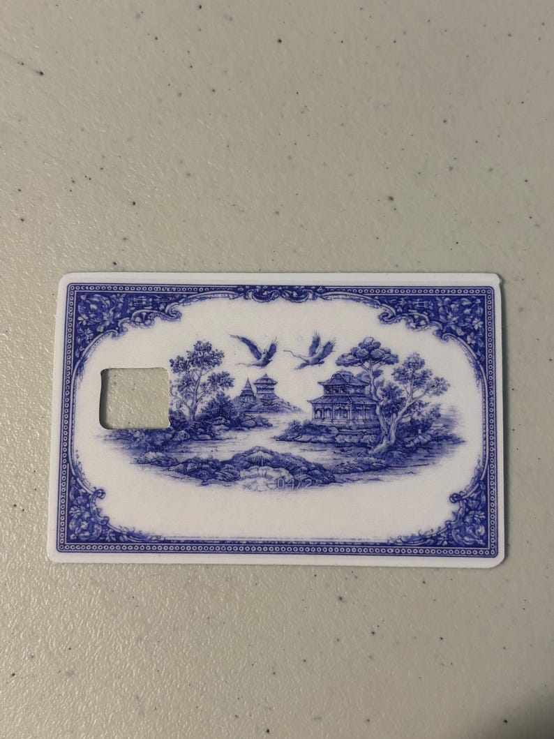 May include: A rectangular, white and blue card with a decorative design. The blue design features a landscape with buildings, trees, and flying birds. The card has a rectangular cutout on the left side.
