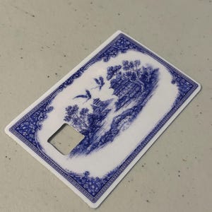 May include: A white and blue bottle opener with a decorative Asian-inspired landscape design. The design features a pagoda, trees, and birds in flight. The opener has a rectangular shape with a square cutout.