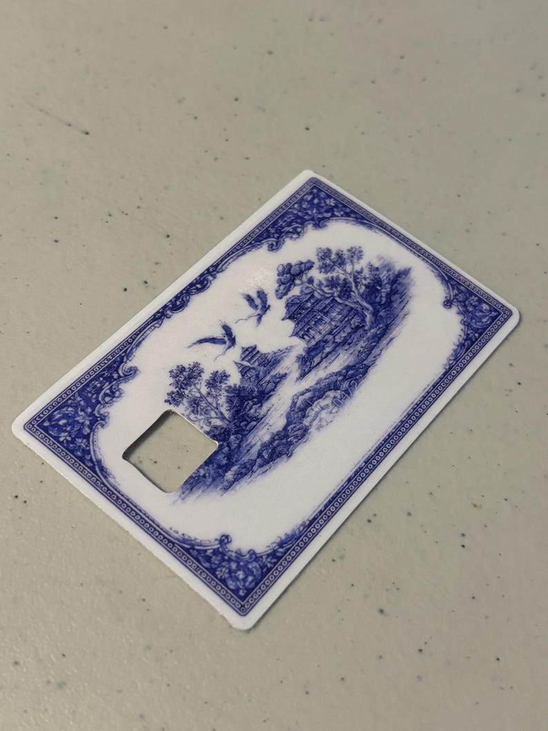 May include: A white and blue bottle opener with a decorative Asian-inspired landscape design. The design features a pagoda, trees, and birds in flight. The opener has a rectangular shape with a square cutout.