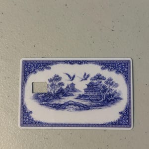 May include: A rectangular card with a blue and white landscape design. The design features a bridge, buildings, trees, and flying birds. The card has a keyhole-shaped cutout on the left side and a decorative border.