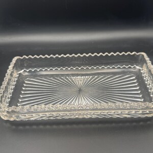 May include: A rectangular clear glass tray featuring a sunburst pattern radiating from the center of the base. The edges of the tray are finished with a decorative scalloped border, and the sides display a subtle geometric textured pattern.