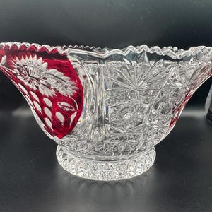 May include: A clear and ruby red cut glass bowl featuring intricate starburst patterns, geometric cross-hatching, and a scalloped rim. The base is circular with a textured diamond pattern, and the body displays alternating clear and red panels.