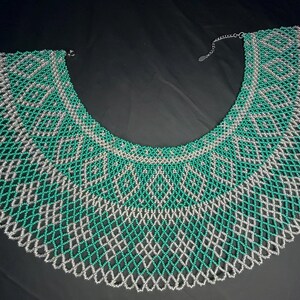 Op de afbeelding: Een statement ketting met een brede kraag. De ketting is gemaakt van groene en zilveren kralen, gerangschikt in een geometrisch patroon. De ketting wordt gepresenteerd op een zwarte achtergrond.