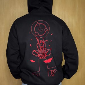 May include: Black hooded sweatshirt with a red graphic on the back. The graphic features a character with a sword and a circular design with three tomoe. The sweatshirt has a drawstring hood and long sleeves.