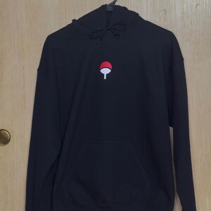 May include: Black hoodie with a front pocket and a drawstring hood. The hoodie features a small embroidered design of a red circle with a white fan-like shape in the center. The hoodie is hanging on a black hanger.
