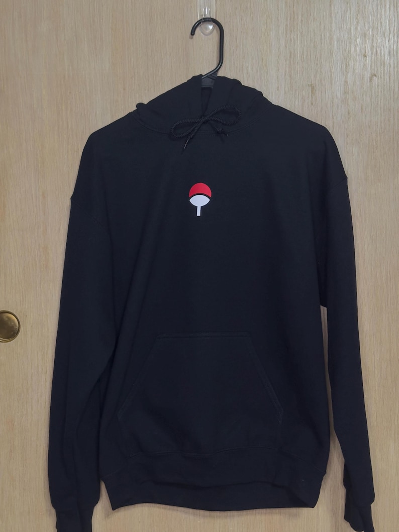 May include: Black hoodie with a front pocket and a drawstring hood. The hoodie features a small embroidered design of a red circle with a white fan-like shape in the center. The hoodie is hanging on a black hanger.