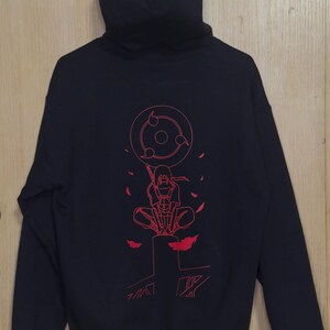 May include: Black hooded sweatshirt with a red graphic on the back. The graphic features a character with a circular design and other elements. The sweatshirt has a hood and long sleeves.