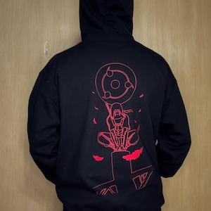 May include: Black hooded sweatshirt with a red graphic on the back. The graphic features a character with a sword, surrounded by a circular design and stylized elements. The sweatshirt has a drawstring hood.