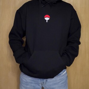 May include: Black hooded sweatshirt with a kangaroo pocket and a drawstring. The sweatshirt features a small red and white emblem on the chest. The person is wearing blue jeans.