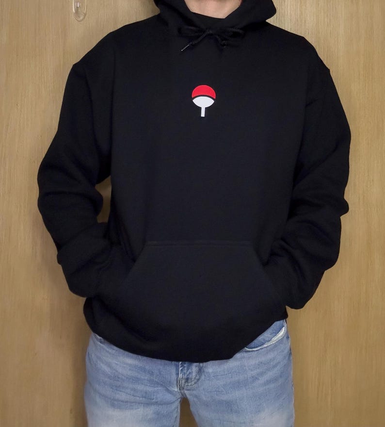 May include: Black hooded sweatshirt with a kangaroo pocket and a drawstring. The sweatshirt features a small red and white emblem on the chest. The person is wearing blue jeans.