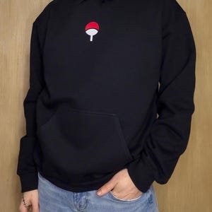 May include: Black hooded sweatshirt with a red and white emblem on the chest. The sweatshirt has a front pocket and a drawstring hood. The person is wearing blue jeans. The emblem is a stylized design.