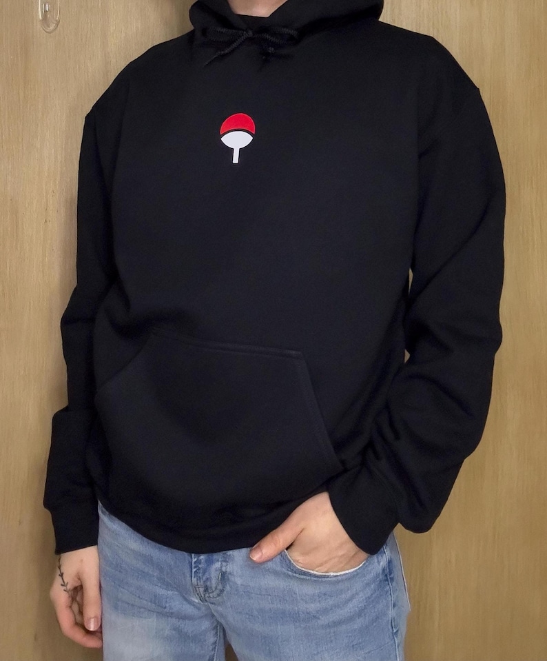 May include: Black hooded sweatshirt with a red and white emblem on the chest. The sweatshirt has a front pocket and a drawstring hood. The person is wearing blue jeans. The emblem is a stylized design.