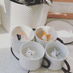 Op de afbeelding: Vier witte keramische mokken met zwarte handvatten en kattenbeeldjes aan de binnenkant. Elke mok staat op een bijpassende schotel met een zwarte rand. De kattenbeeldjes zijn in verschillende kleuren, waaronder oranje, grijs en lapjeskat. Een witte, getextureerde pot met een plant staat op de achtergrond.
