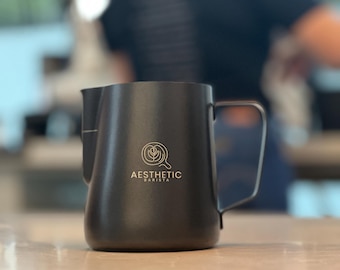 May include: A matte black milk frothing pitcher with a handle and a spout, sitting on a light-colored surface. The pitcher has the words "Aesthetic Barista" printed on the front, along with a coffee bean logo.