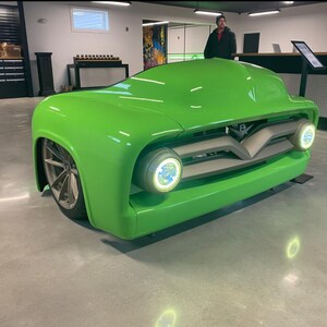 May include: A custom, bright green car with a unique design. The vehicle features a streamlined body, large front headlights, and a silver grill. The car is displayed indoors on a concrete floor. The wheels are silver.