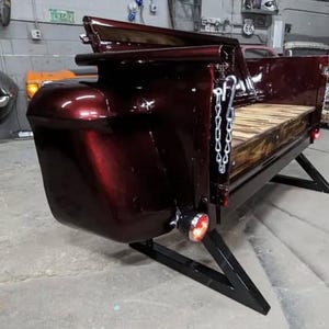 May include: A custom-built burgundy truck bed, mounted on a black metal frame. The bed features a wooden floor and is equipped with taillights and a chain. The design is reminiscent of classic automotive styling.