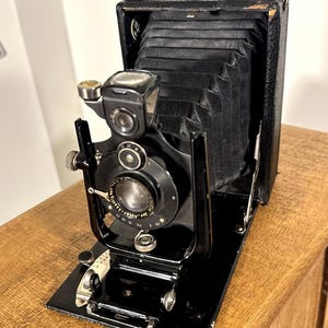 May include: A vintage black folding camera with a leather handle and bellows. The camera has a lens, viewfinder, and various dials and levers. It is set on a black base and appears to be in good condition. The camera is likely from the early to mid-20th century.