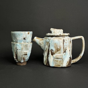Op de afbeelding: Een keramische theepot met deksel en twee gestapelde keramische kopjes, allemaal met een getextureerd, ongelijkmatig oppervlak met een lichtblauwe glazuur over een donkerbruine basis, met een handgevormde, organische vorm en kleidetails.