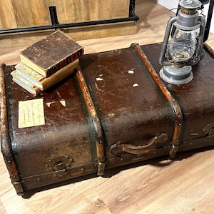 May include: A weathered dark brown vintage trunk with metal latches and leather handle, featuring a stack of two aged hardcover books and a silver-toned metal oil lantern placed on top. A small rectangular paper label is attached to the trunk surface.