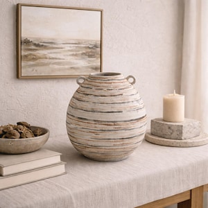 Peut inclure: Un grand vase en céramique rayé avec deux poignées est posé sur une table. Le vase présente des bandes horizontales blanches, marron et grises. Une peinture de paysage encadrée, un bol d'objets décoratifs, une bougie et des livres sont également sur la table.