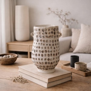 Peut inclure: Un vase en céramique blanc cassé texturé, de forme cylindrique, avec deux petites anses. Le vase est orné d'un motif de creux circulaires bruns. Il est posé sur une pile de livres, suggérant un élément de décoration intérieure.